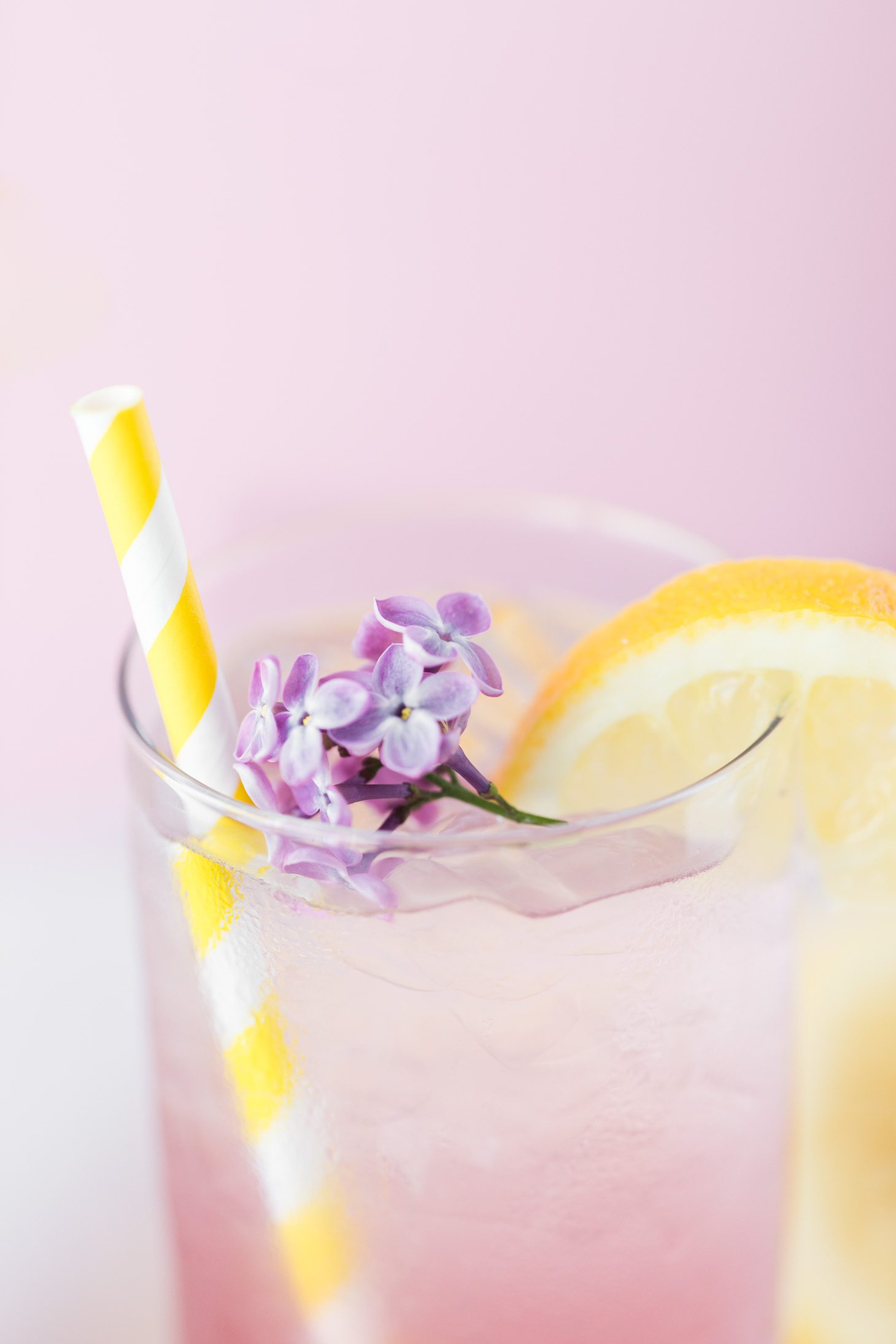 Glass of Lilac Lemonade with yellow striped straw, lemon slice and lilac blossoms. 