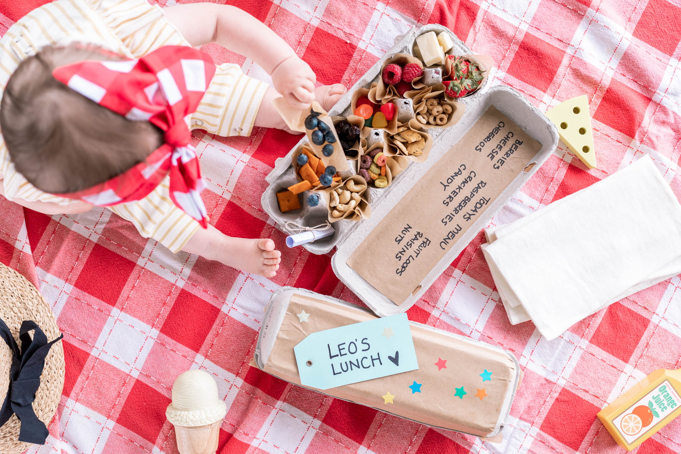 Overhead view of Jillian Harris' daughter Annie sitting on a red plaid cloth with snacks at a Picnic.