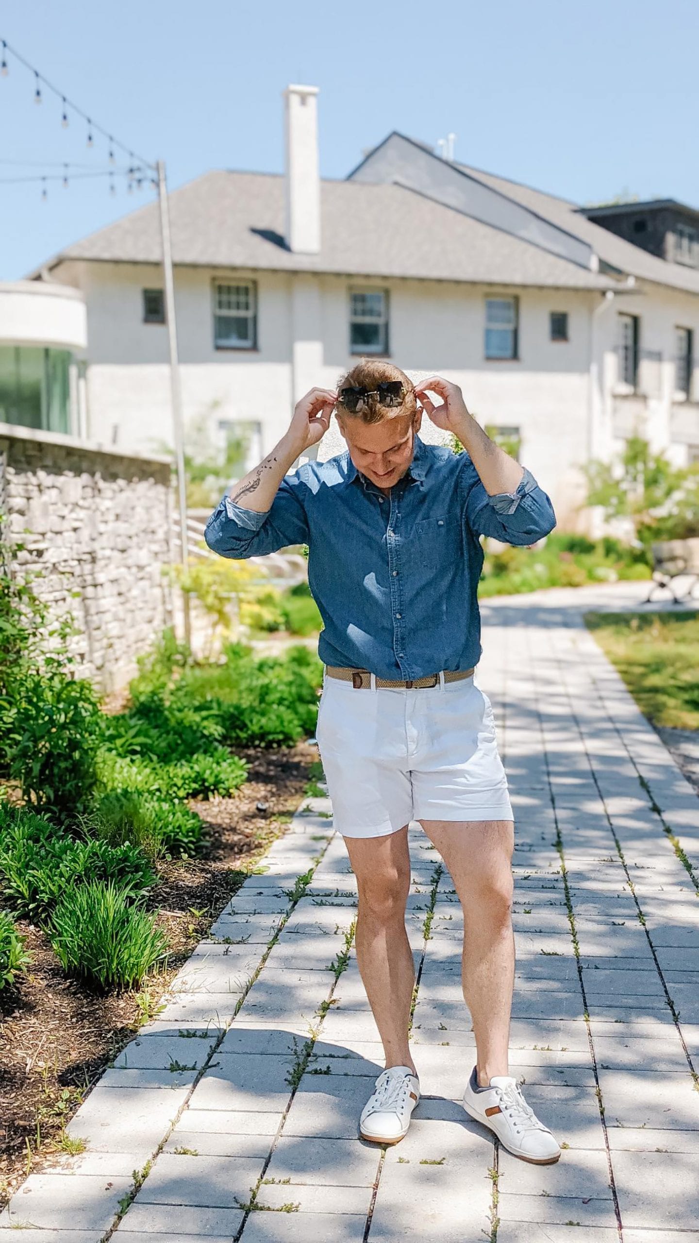 Christopher standing outside on a stone path wearing white shorts and a denim button down shirt, one of his 2021 Summer Outfits.