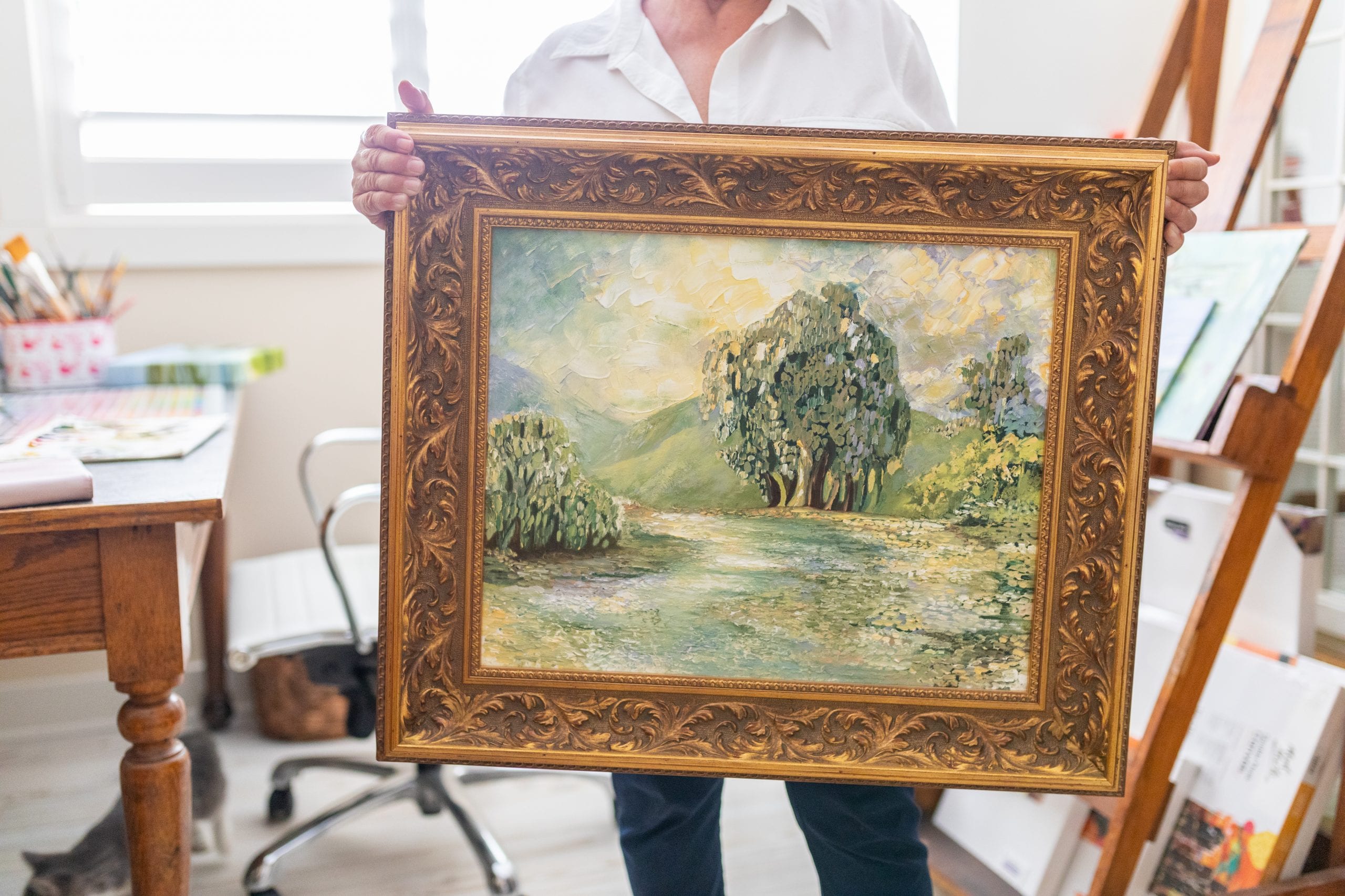 Peggy Harris holding a framed painting of an outdoor scene with trees, a field and a rolling green hill in the background.