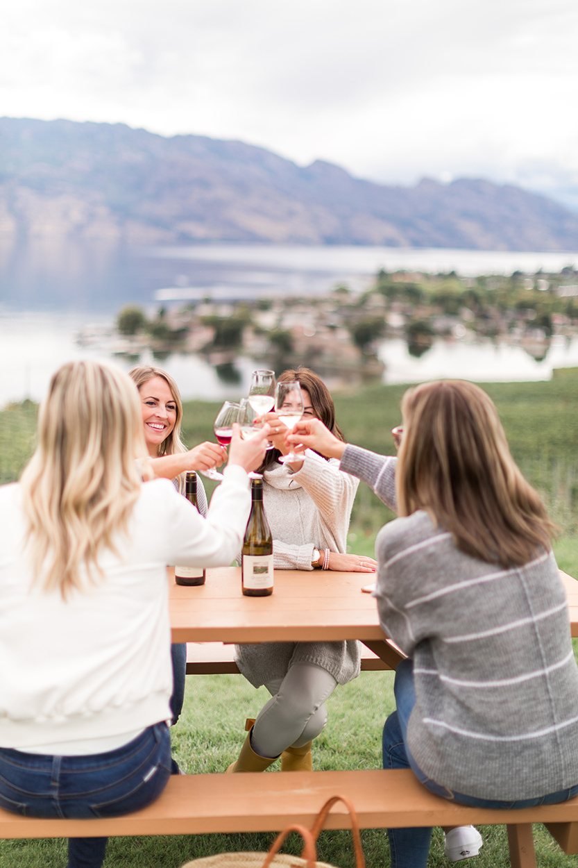 Jillian Harris and a few members from Team Jilly celebrating at Quail's Gate. They are sitting at a picnic table and doing a cheers with their wine glasses. 