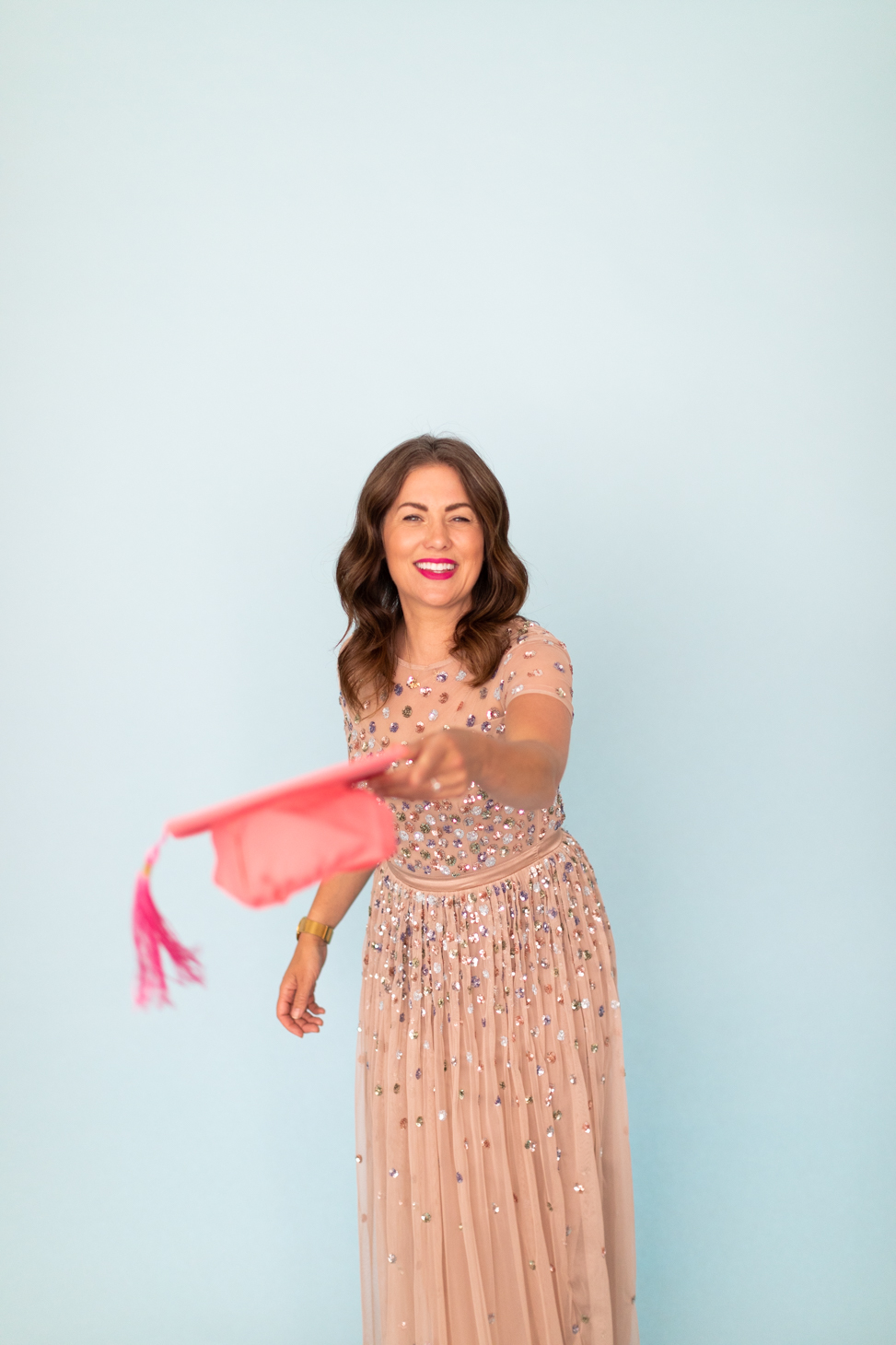 Jillian Harris against a blue backdrop holding a pink graduation cap wearing a pink sparkly dress to celebrate one year of The Jilly Academy.