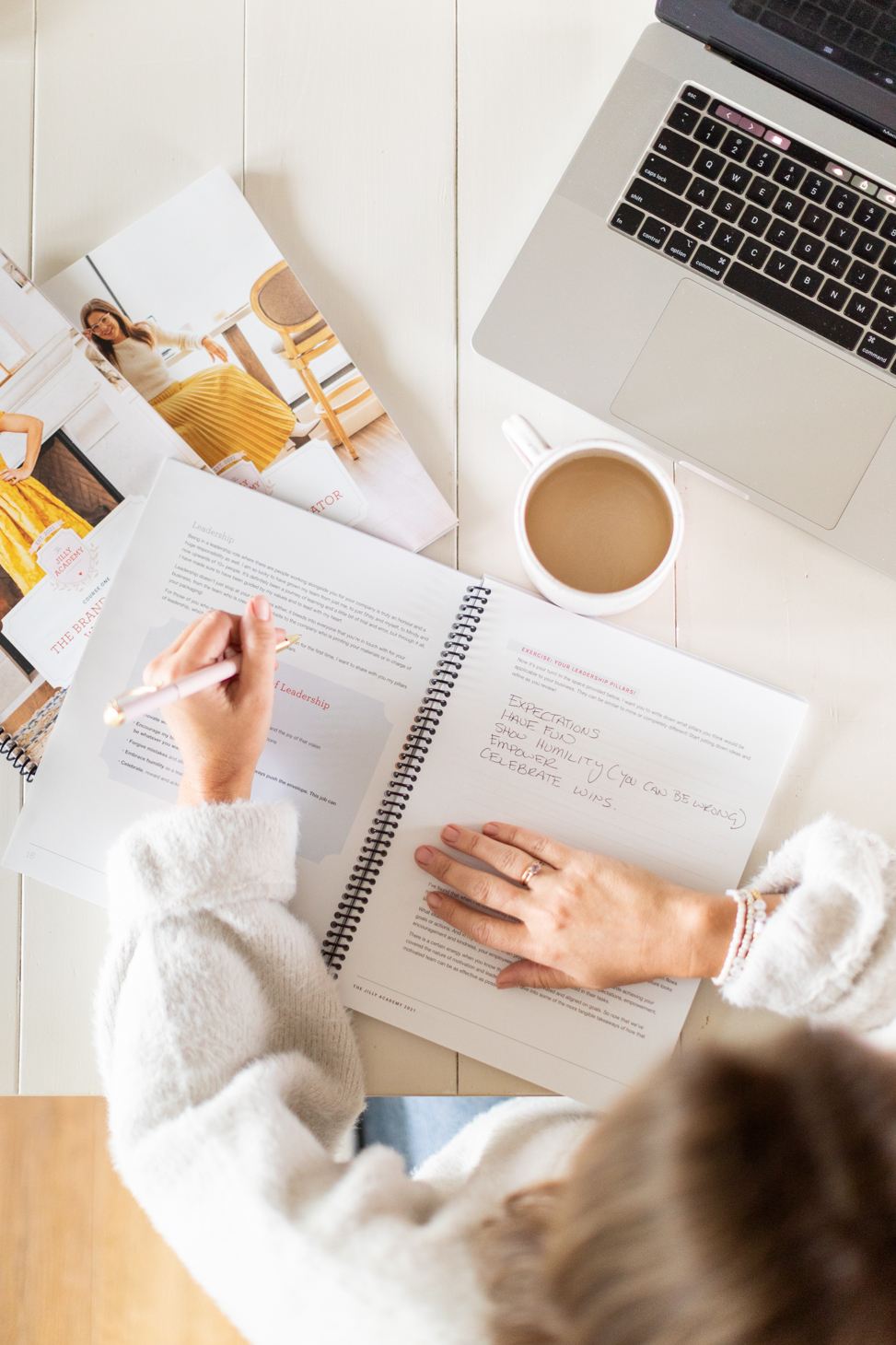 Overhead shot of Jillian Harris showcasing The Jilly Academy Workbooks with a laptop and coffee mug on the table in honour of our one year celebration. 