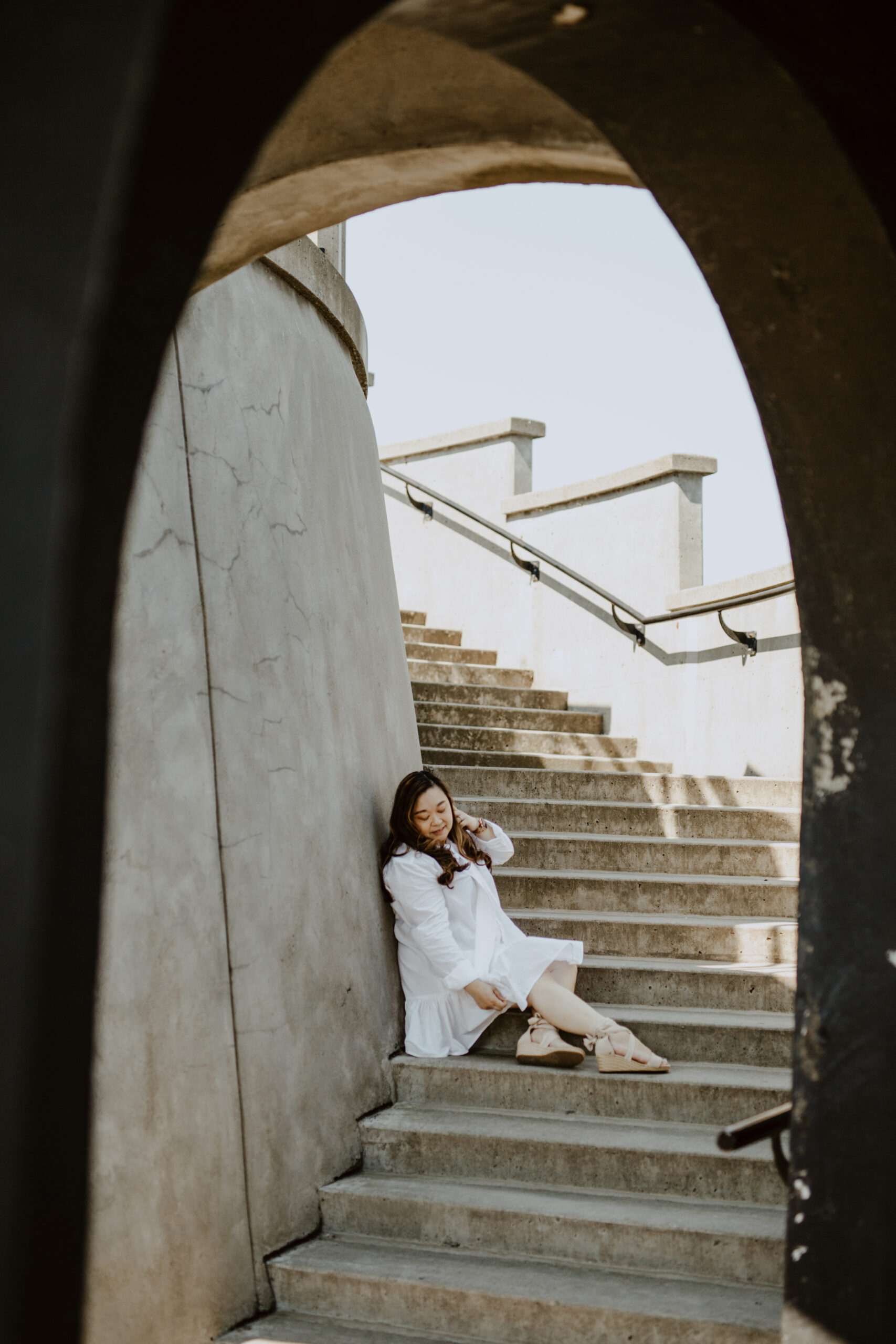 Anne Mok of Purpose in View/ Alumni to The Jilly Academy sitting on a concrete staircase wearing a white dress and beige wedges. 