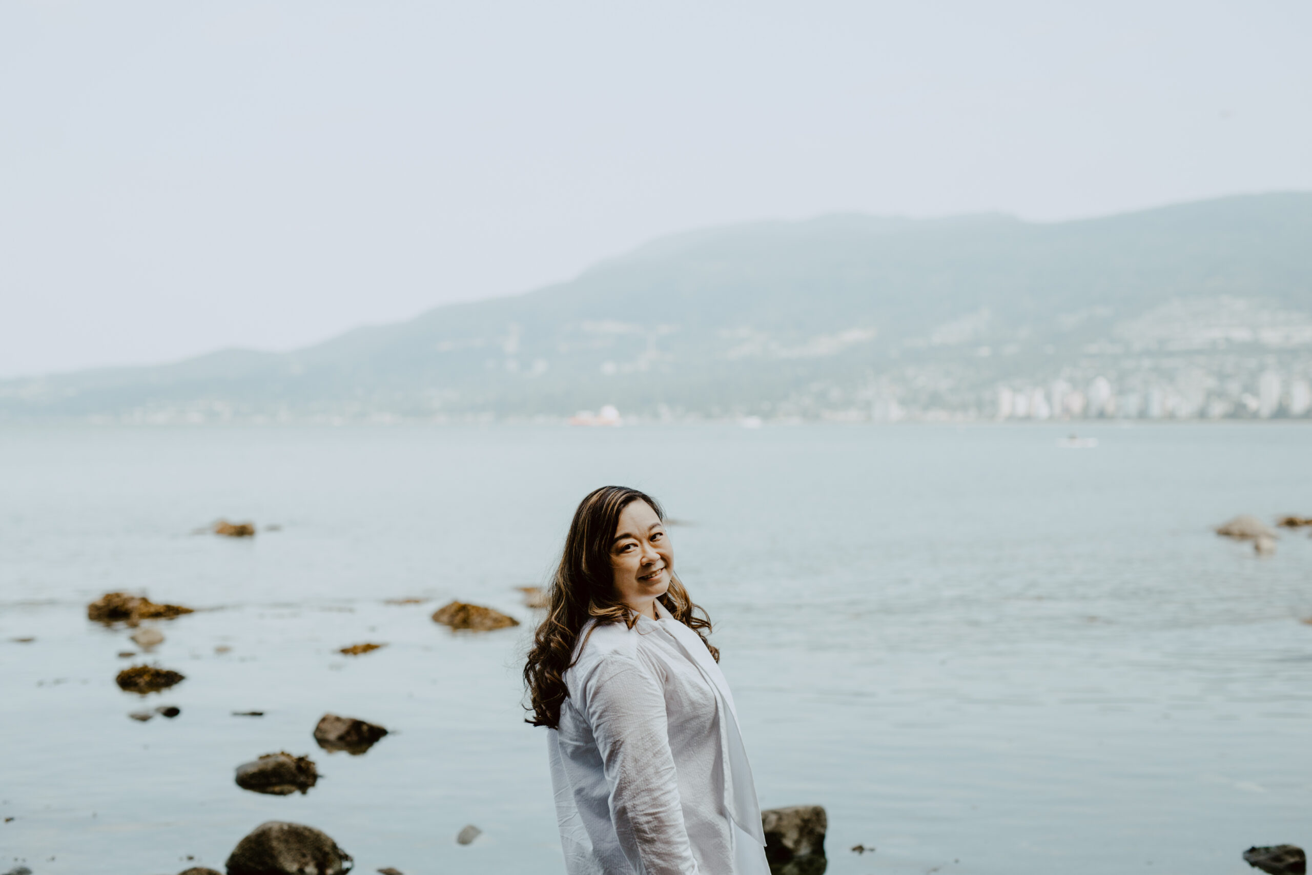 Anne Mok of Purpose in View/ Alumni to The Jilly Academy, looking over her shoulder wearing a white dress standing in front of a mountain and lake landscape. 