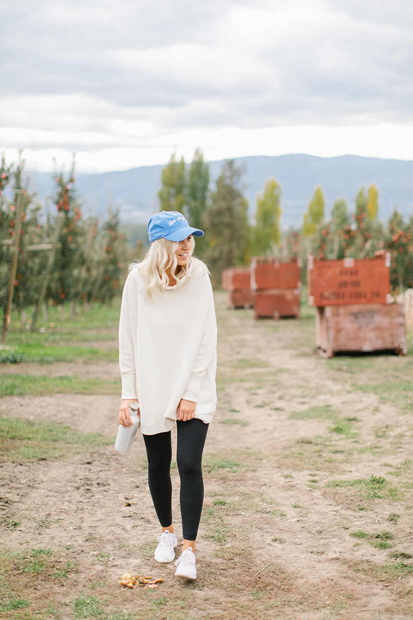 Shay of Team Jilly walking through an orchard wearing sneakers, leggings, sweater and a blue hat holding a water bottle as part of her morning habits. 