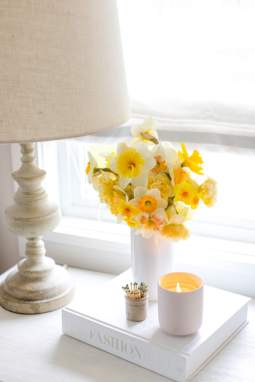 Jillian Harris' nightstand with a lamp, a lit candle, a jar of matches, and a vase of daffodils on top of a coffee table book to set the scene for good morning habits. 