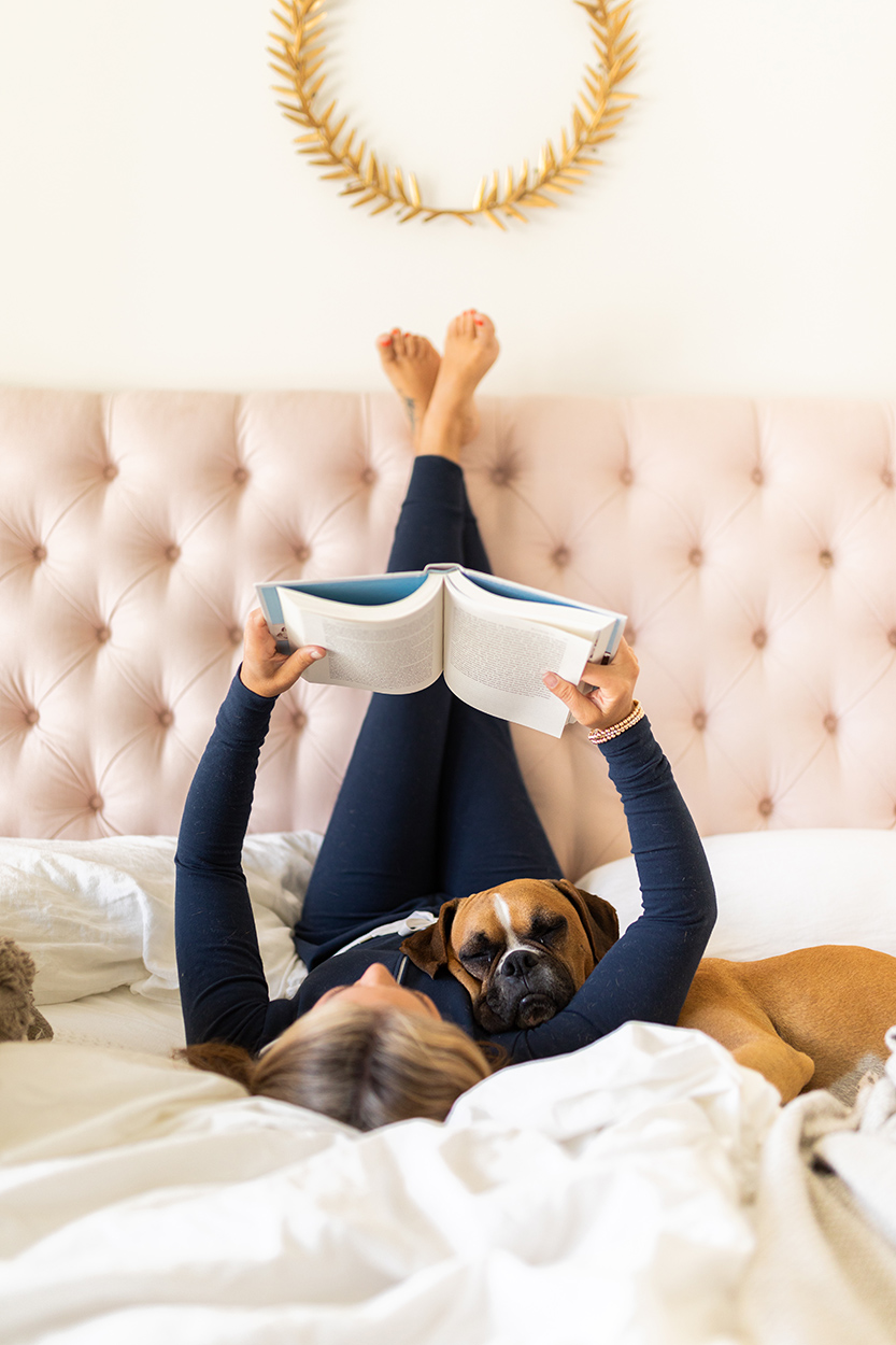 Jillian Harris laying on her bed with her feet up on the headboard wearing a navy Smash and Tess Shay Romper reading a book while cuddling her boxer dog Nacho as part of her morning habits.