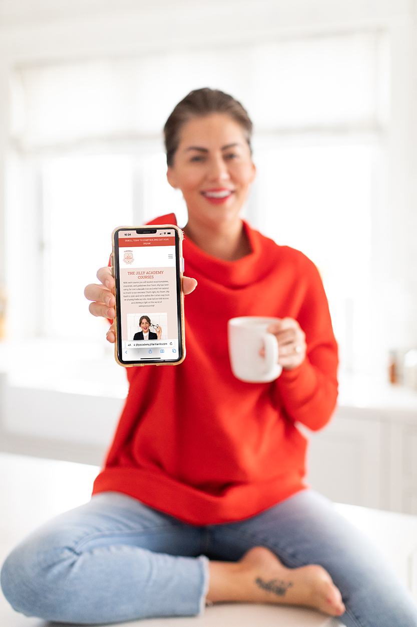 Jillian Harris sitting on her kitchen counter with showing her phone with The Jilly Academy website home page for the example of owned platforms.