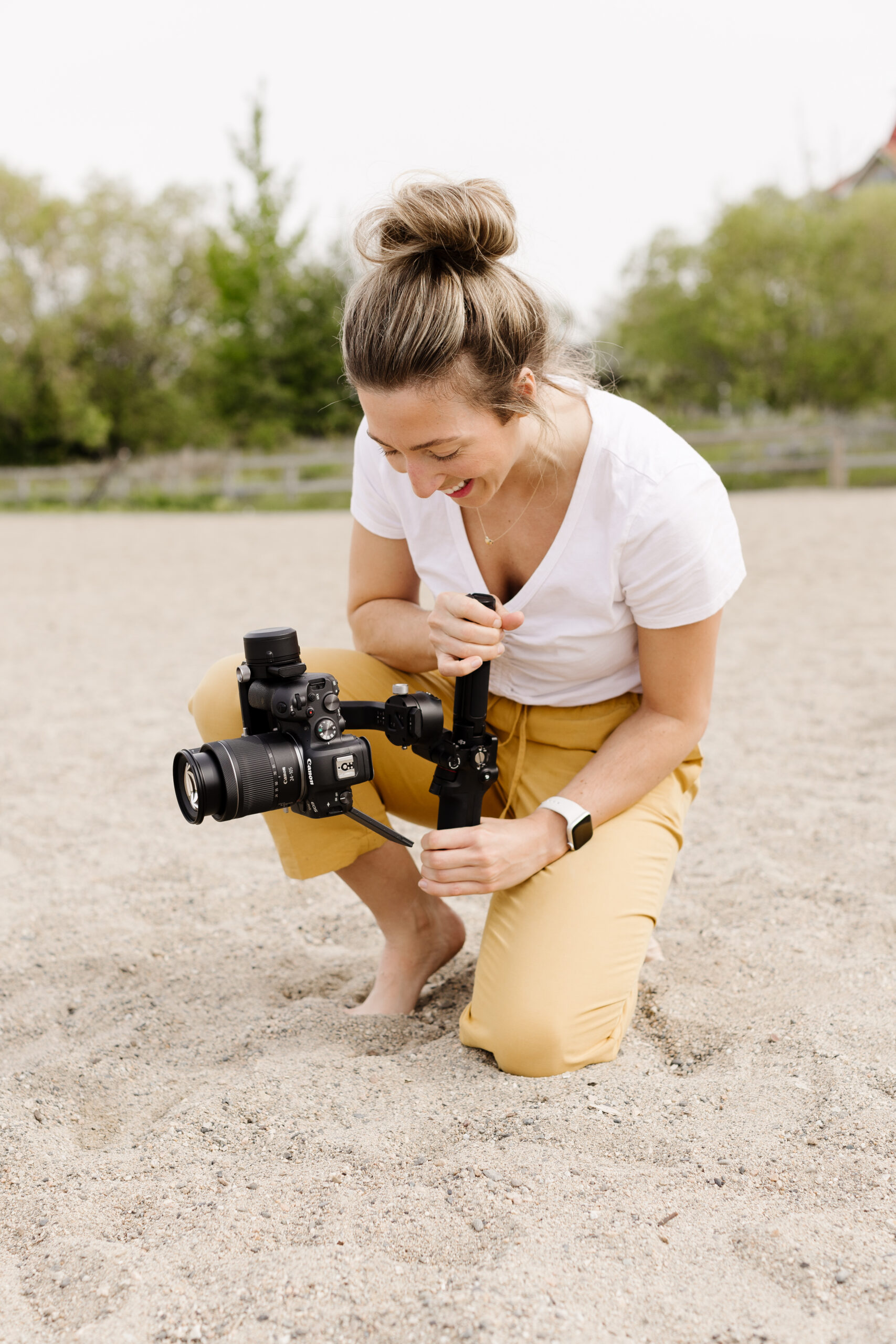 Maggie of Team Jilly on an Okanagan beach wearing a white t-shirt and yellow pants holding her camera to take video at a photoshoot. 