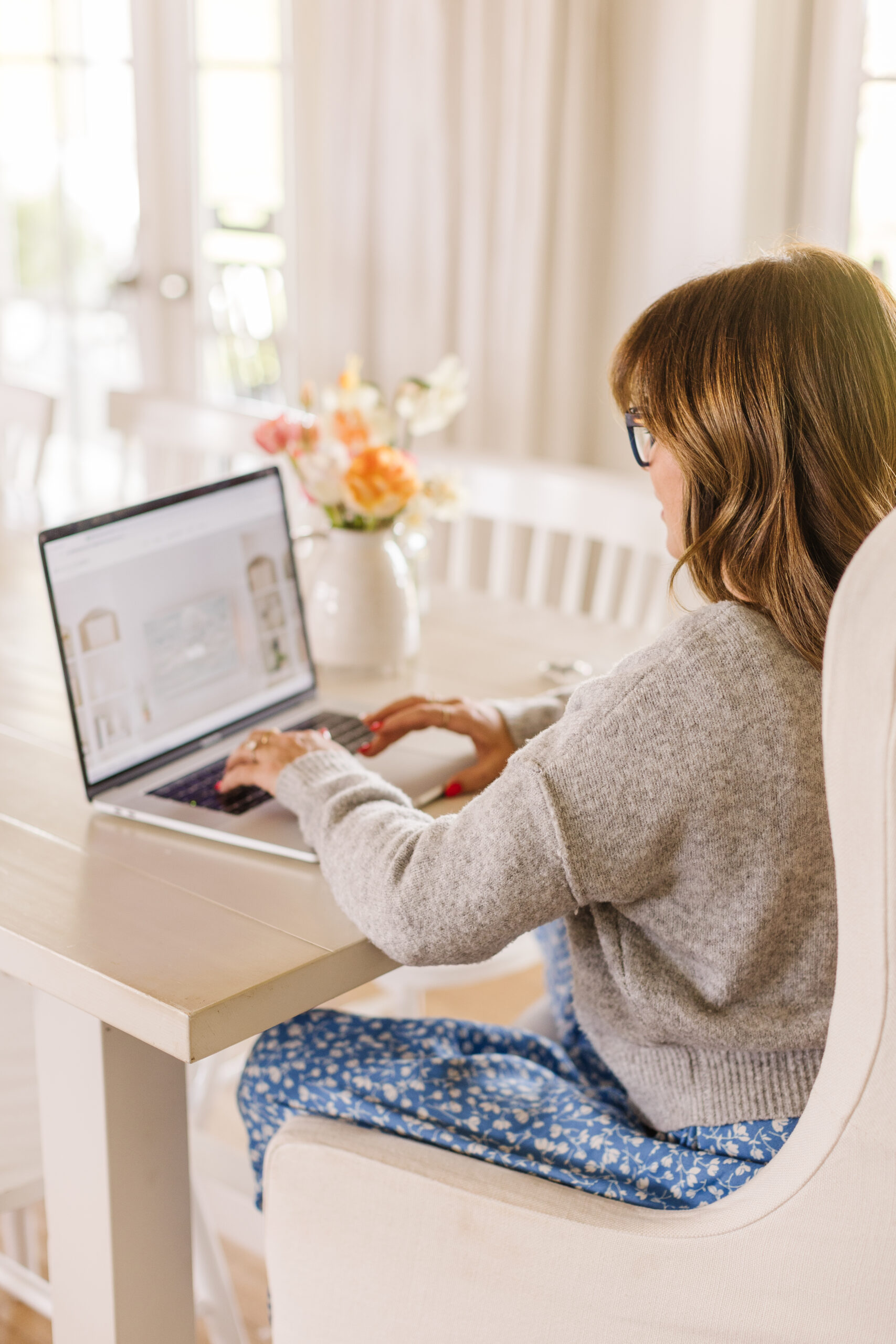 Overview of Jillian Harris sitting at her dining room table wearing a blue dress with white flowers and a grey cardigan working on her laptop to start her video content strategy for the week. 