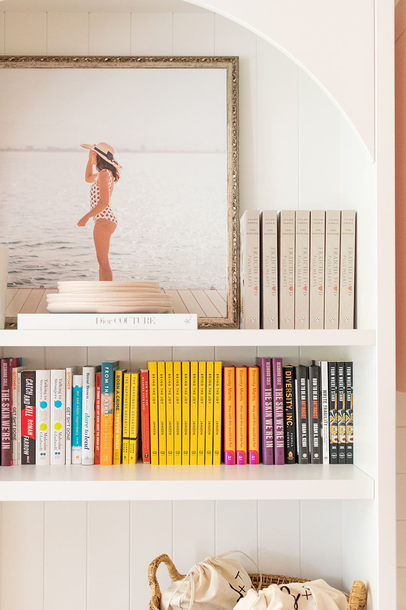 Jillian Harris' office library shelf showing some must have reads. Shelves are decorated with a photo of Jill in a one piece bathing suit wearing a beach hat, Fraiche Food Full Hearts cookbooks and a stack of plates.