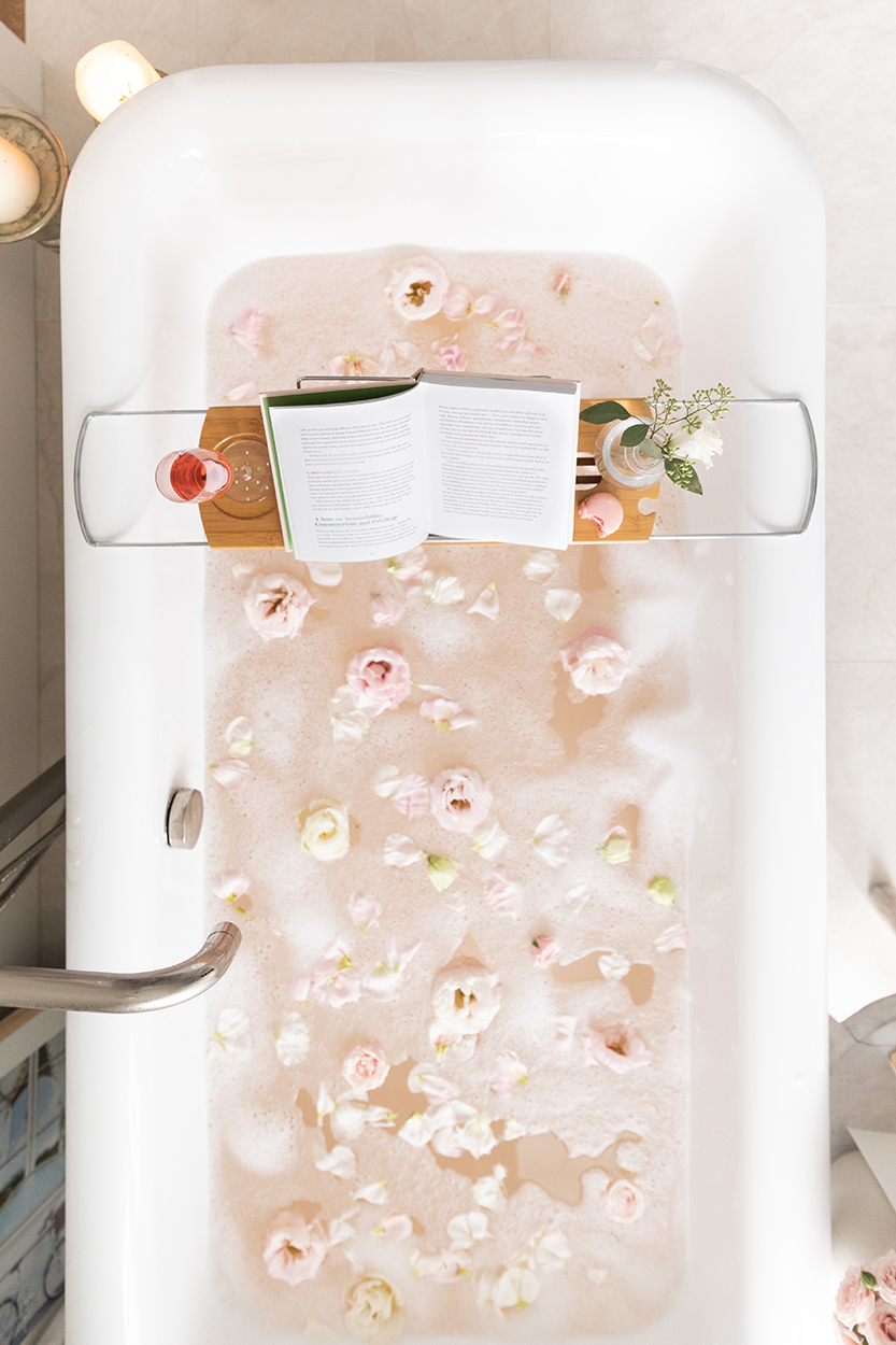 Overview of Jillian Harris bathtub with a must read book on top of a shelf with lots of flower petals as a nice space to curl up and read a book. 