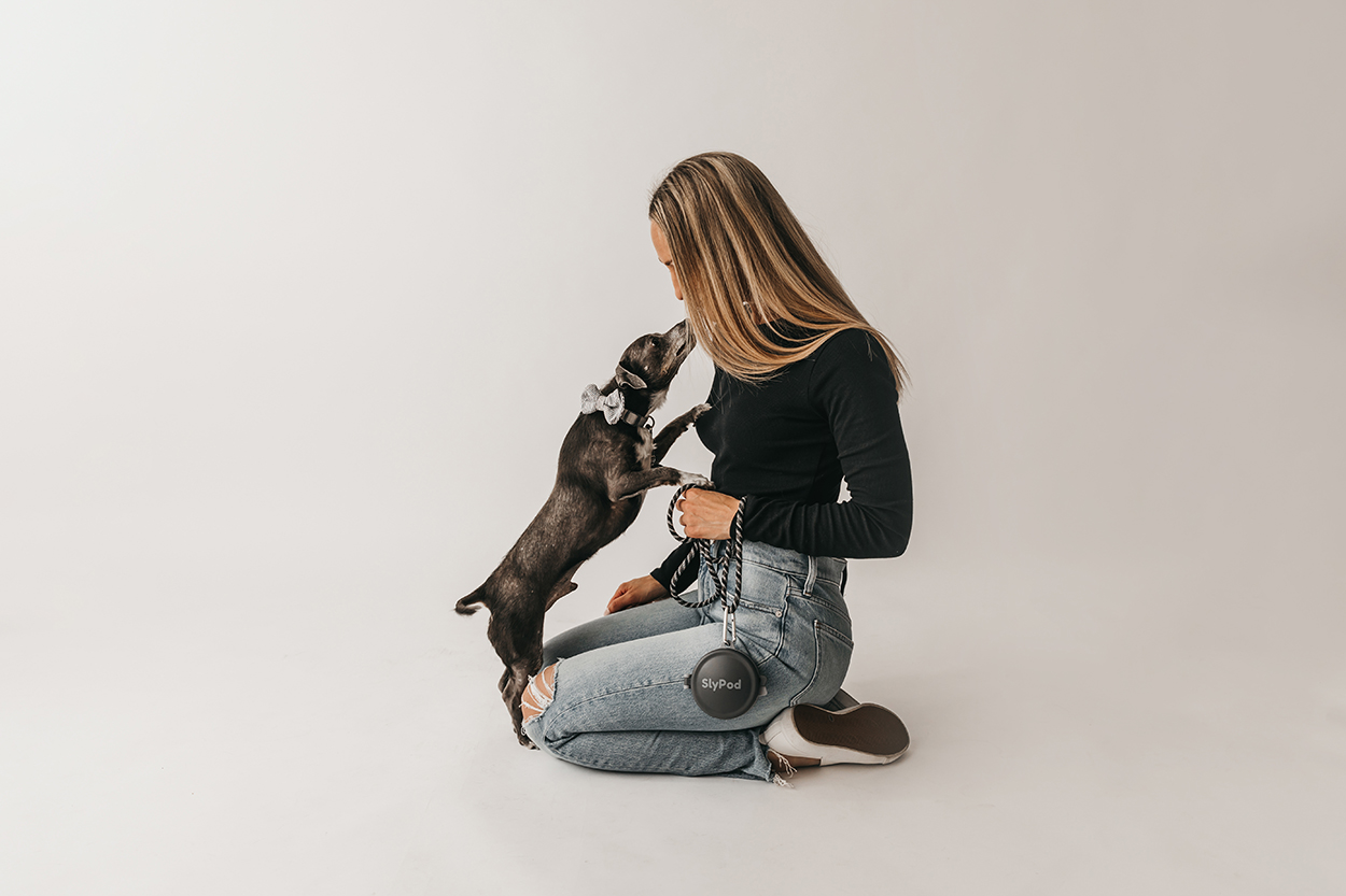 Jilly Academy Alumni member Nicole Duff standing on a white background wearing blue jeans, a black sweater and white sneakers. She is leaning down to kiss her black dog. They are showcasing her product where she took one of her big risks on, SlyPod.