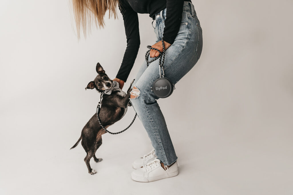 Jilly Academy Alumni member Nicole Duff standing on a white background wearing blue jeans, a black sweater and white sneakers. She is leaning down to pet her black dog. They are showcasing her product where she took one of her big risks on, SlyPod.