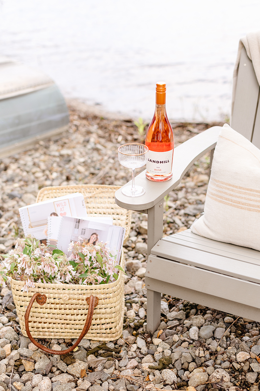 The Jilly Academy workbooks in a basket with flowers next to a Muskoka chair with a wine bottle sitting on the arm rest with a wine glass showcasing the result of pitching a brand partnership. 