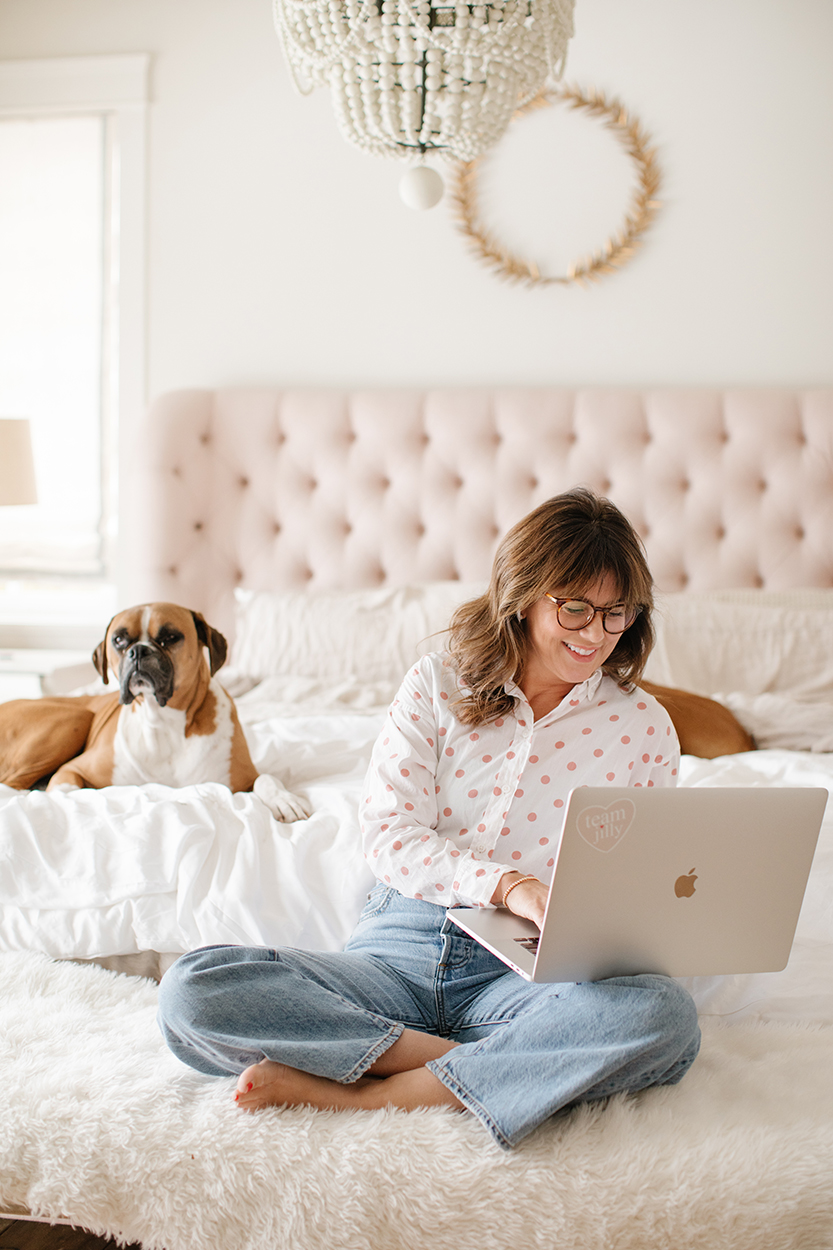 Jillian Harris sitting on her bed wearing a pink and white polkadot button up, blue jeans and holding a laptop. Nacho and Peaches, her two Boxer dogs sitting behind her, ready to support restart at any age. 