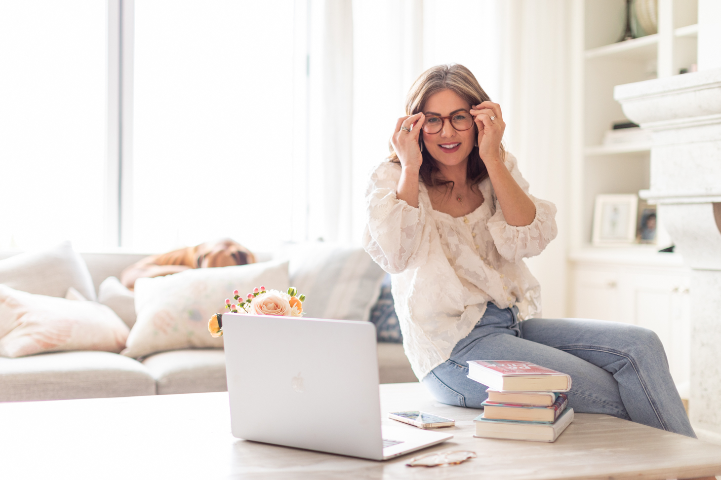 Jillian Harris sitting on her coffee table with a stack of books and laptop putting her glasses on wearing a white top and blue jeans here to support you pitching a brand.