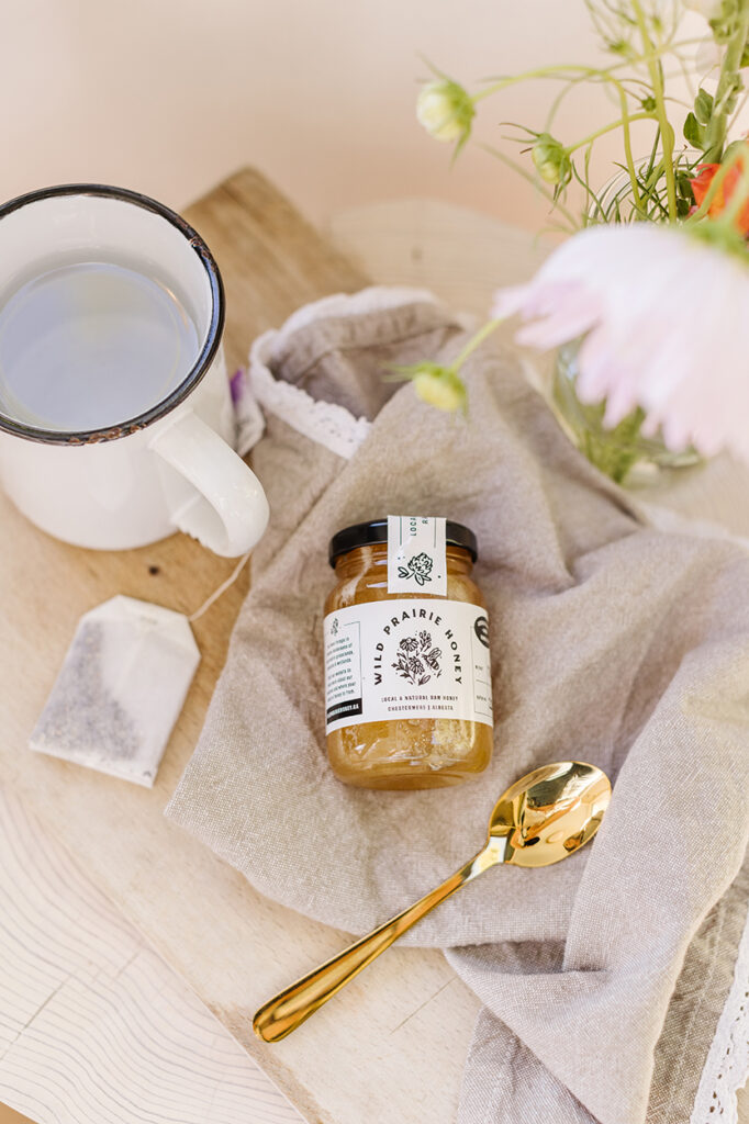 Wild Prairie Honey jar is part of the git bag, shown here laying flat on a tea towel next to a spoon, mug, tea bag and flowers.