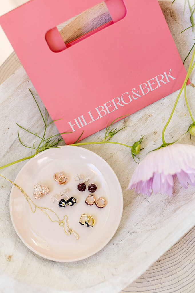 Hillberg and Berk gift bag with a plate of earrings and a necklace chain on top of a wooden platter and flowers. 