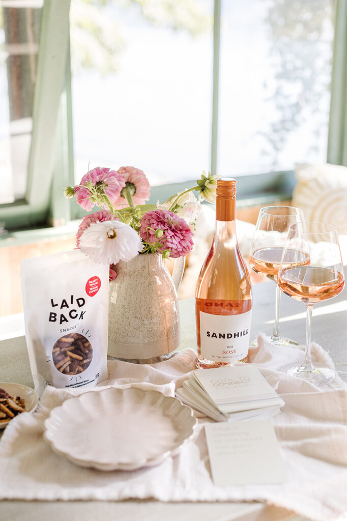 Items in gift bag on farmhouse table: laidback snacks next to a vase of flowers, Sandhill rose with two glasses, Cool to connect cards. 