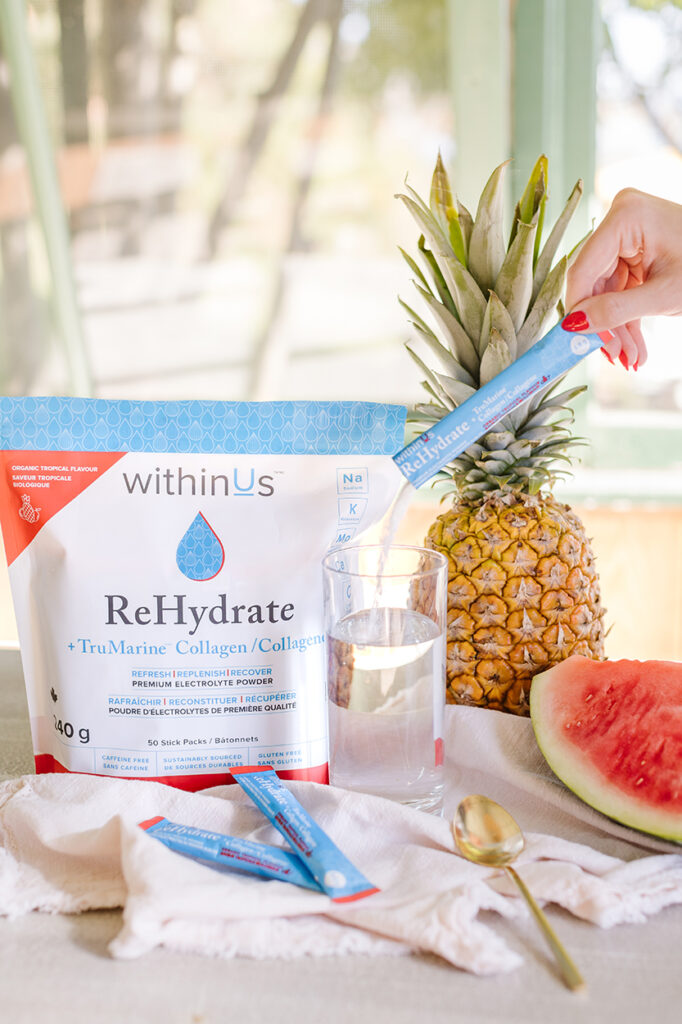 withinUs Rehydrate from the gift bag next to a pineapple, watermelon, glass of water with ReHydrate getting poured in and a spoon. 