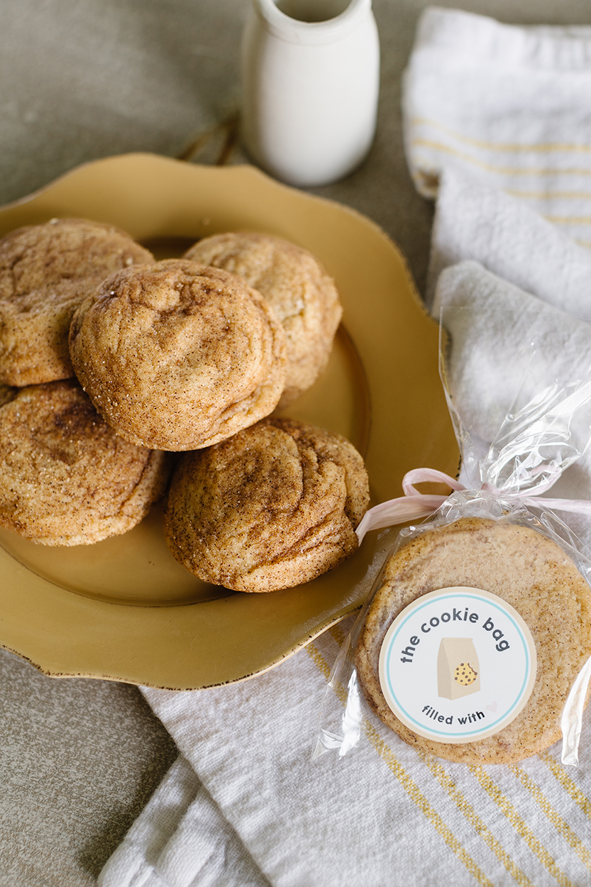 The Cookie Bag featured in the gift bag - handmade cinnamon swirl flavoured cookies on a plate with a milk jug next to it and a tea towel. One cookie is wrapped up with The Cookie Bag logo sticker.