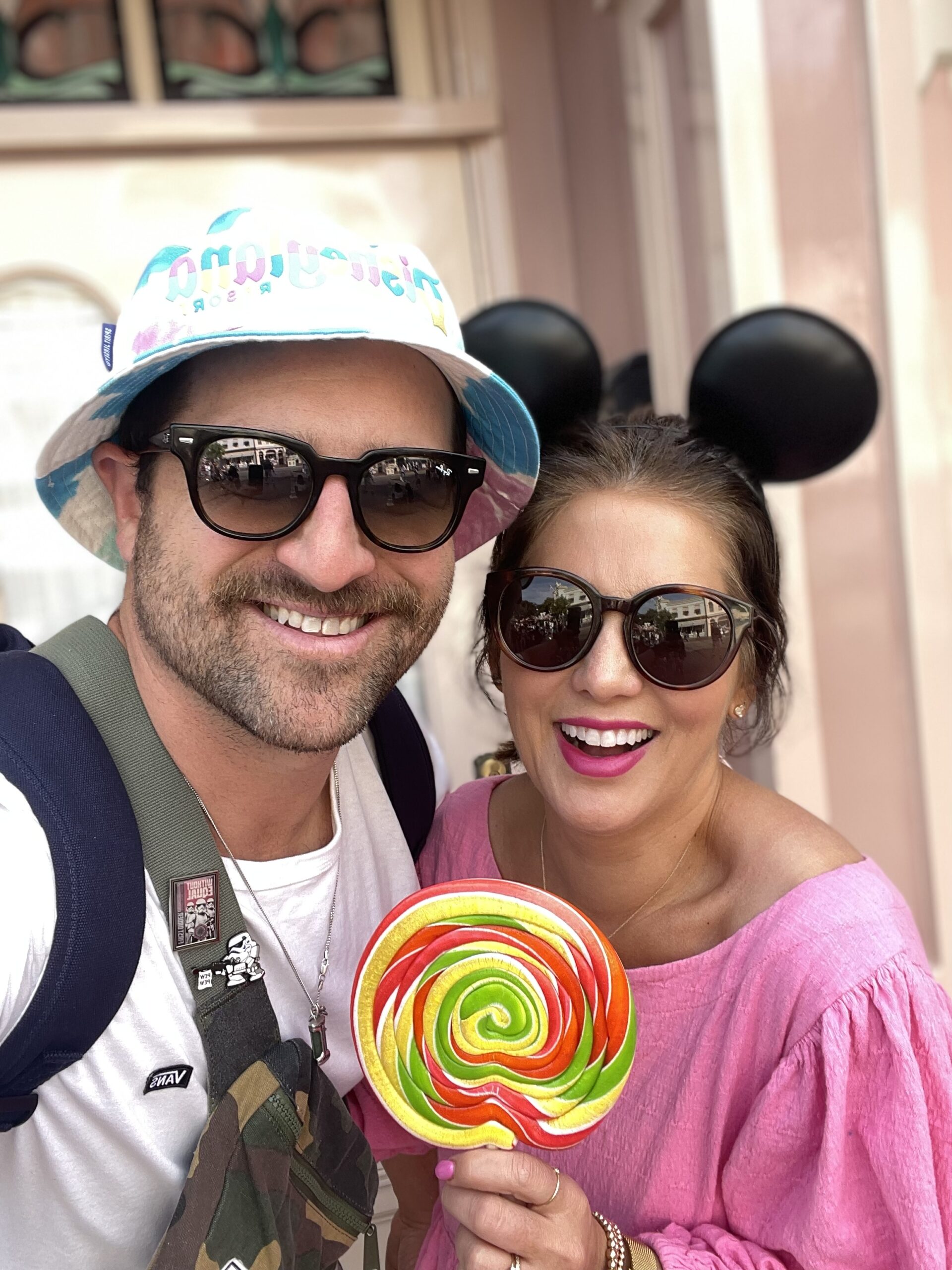 Jillian Harris and Justin Pasutto taking a selfie wearing sunglasses, Disneyland headgear and Jillian holding a large lollypop.