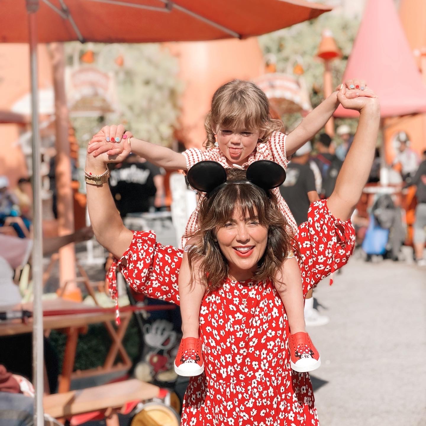 Jillian Harris in a red flower print dress, holding daughter annie on her shoulders, sticking out their tongues