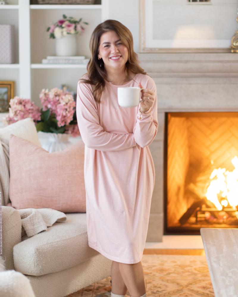 Jillian Harris wearing a sleep dress holding a cup of coffee by a sofa and fireplace. 