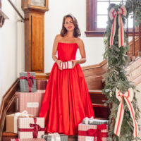 Jillian Harris wearing a festive red dress and standing on a staircase surrounded by The Winter 2025 Jilly Box and Christmas gifts.