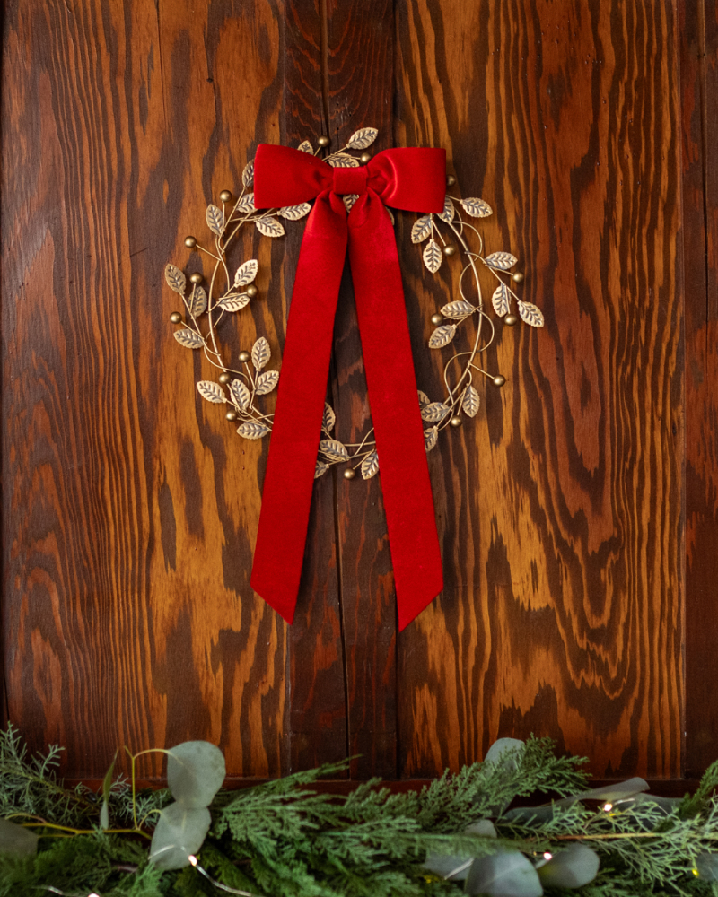 The Jilly Box exclusive Golden Berry Metal Wreath adorned with a red ribbon and hanging on a wooden wall.