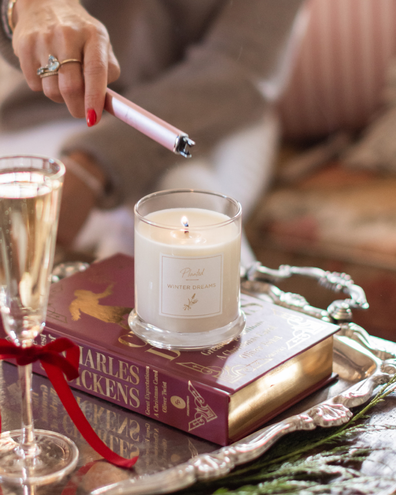 A hand holding a lighter and lighting up the Planted Candle Winter Dreams by a glass of champagne.
